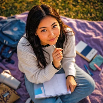 Estudante sentada ao ar livre sobre uma manta, com caderno aberto e caneta na mão, estudando para a redação do ENEM.
